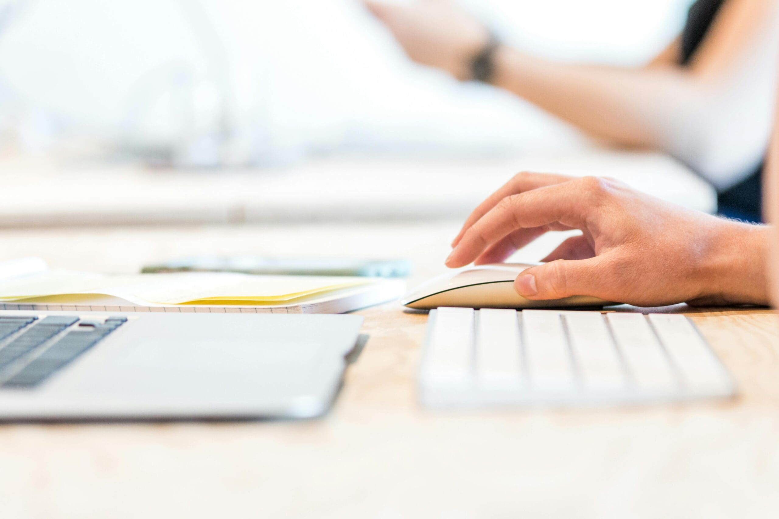 A person's hand using a computer mouse on a wooden desk, with a laptop keyboard visible in the foreground and blurred papers or documents in the background, suggesting a workspace or office environment.