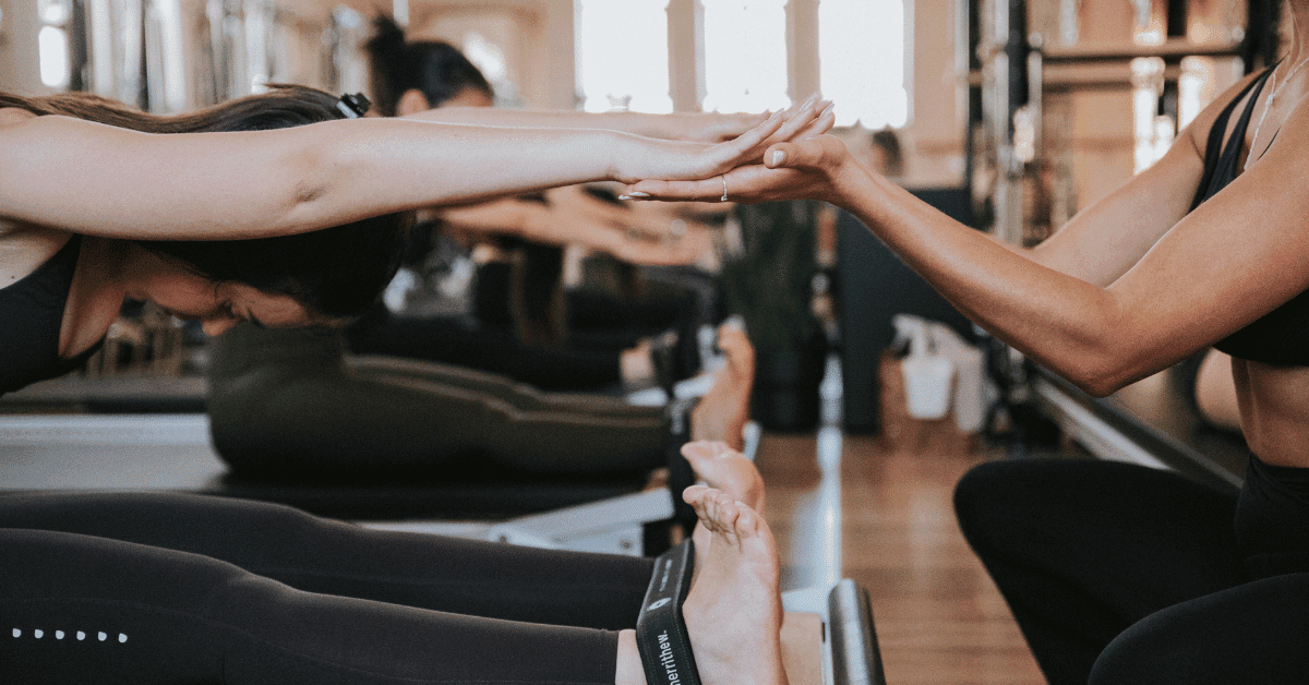 Pilates instructor showing client proper form on reformer in modern studio with laptop displaying website
