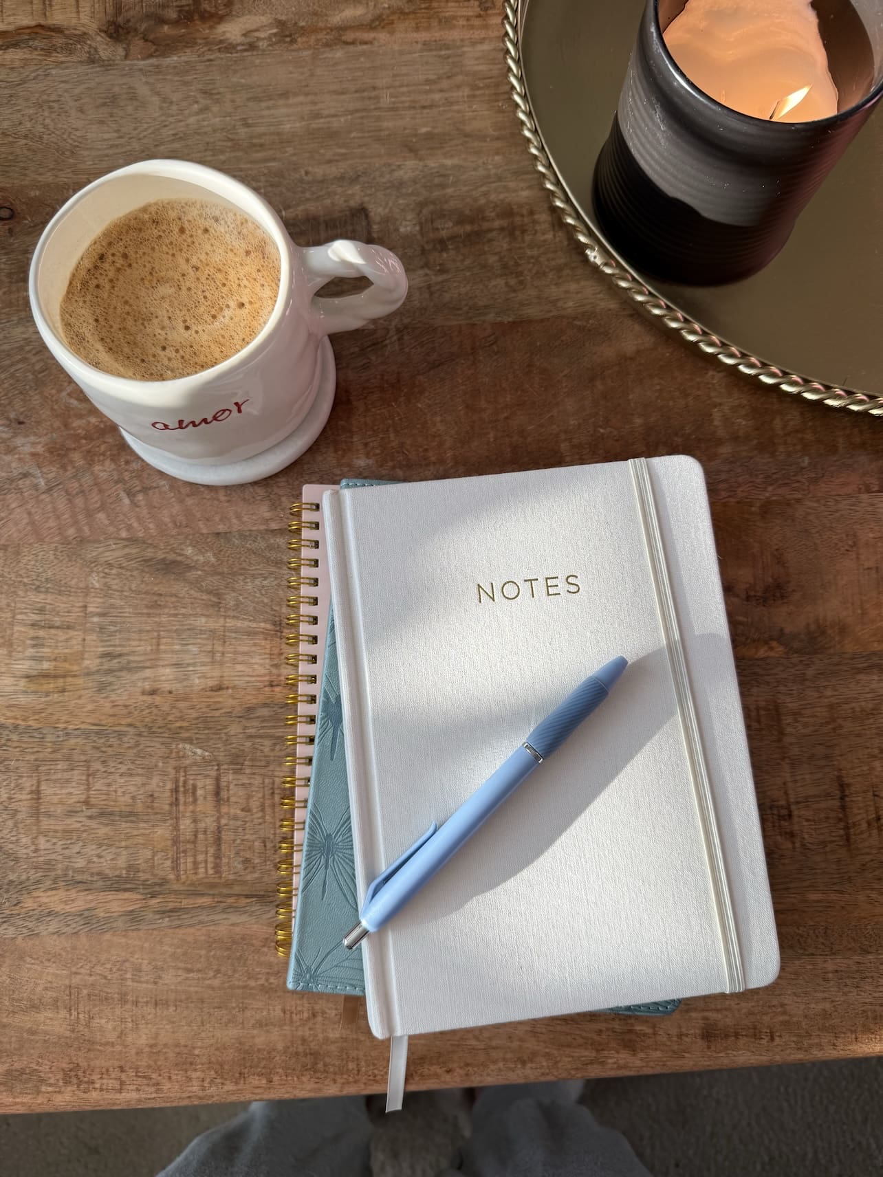 Closed white notebook, with blue pen on top, next to a warm cup of coffee in a white mug on a wooden table.
