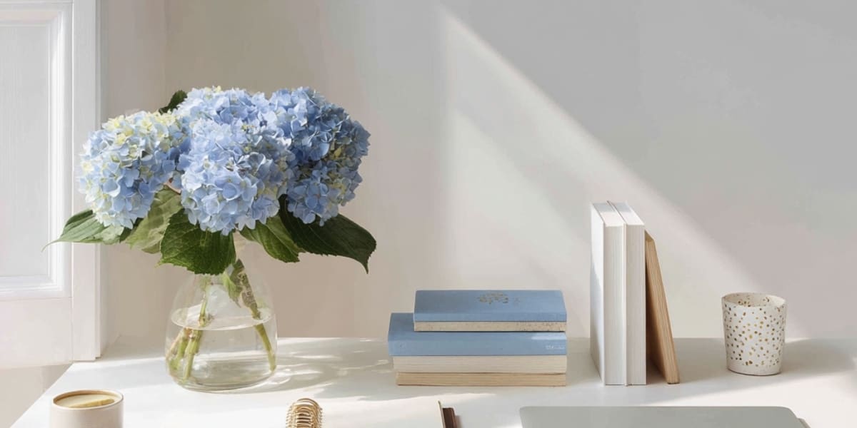 A clean, light-filled desk styled with blue hydrangeas in a glass vase, stacked books, and a speckled candle.